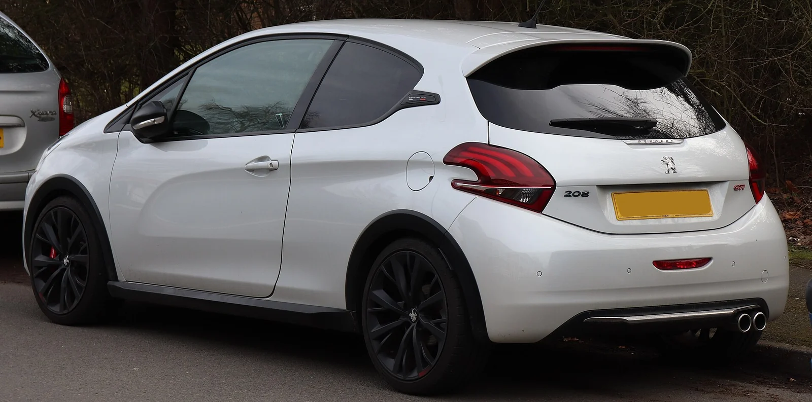 White first-generation Peugeot 208 GTi photographed from the rear three-quarter angle, showing its roof spoiler, black alloy wheels and dual exhaust