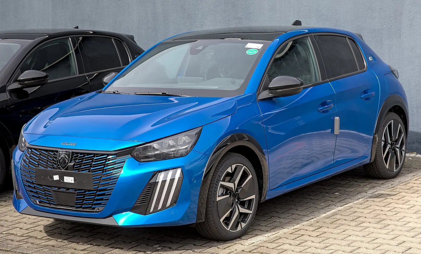 Blue second-generation Peugeot e-208 photographed from the front three-quarter angle, showing its claw-shaped LED daytime running lights and chequered grille pattern