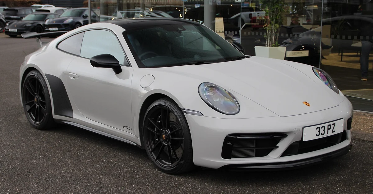 White Porsche 911 992-generation Carrera GTS coupe photographed from the front three-quarter angle outside a dealership, with black wheels and a GTS badge on the front wing