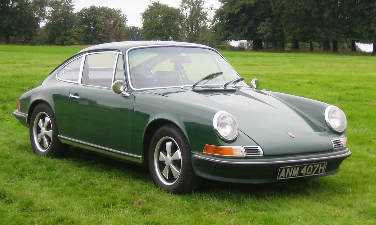 Dark green early Porsche 911 photographed from the front three-quarter angle on grass, with round headlamps and chrome bumpers