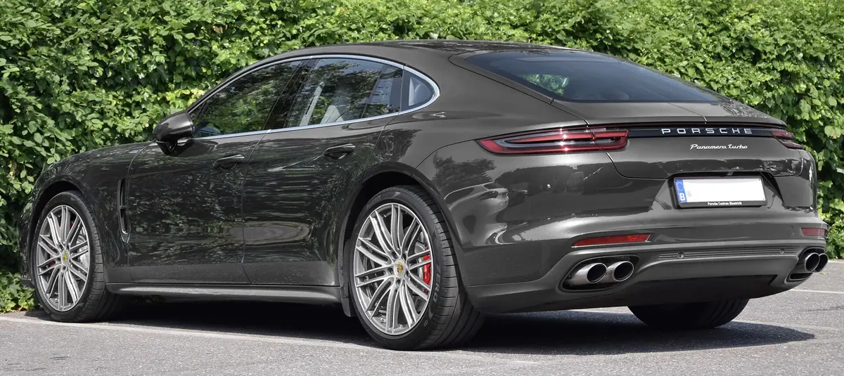 Dark grey Porsche Panamera Turbo liftback photographed from the rear three-quarter angle beside a hedge, with a full-width LED light bar