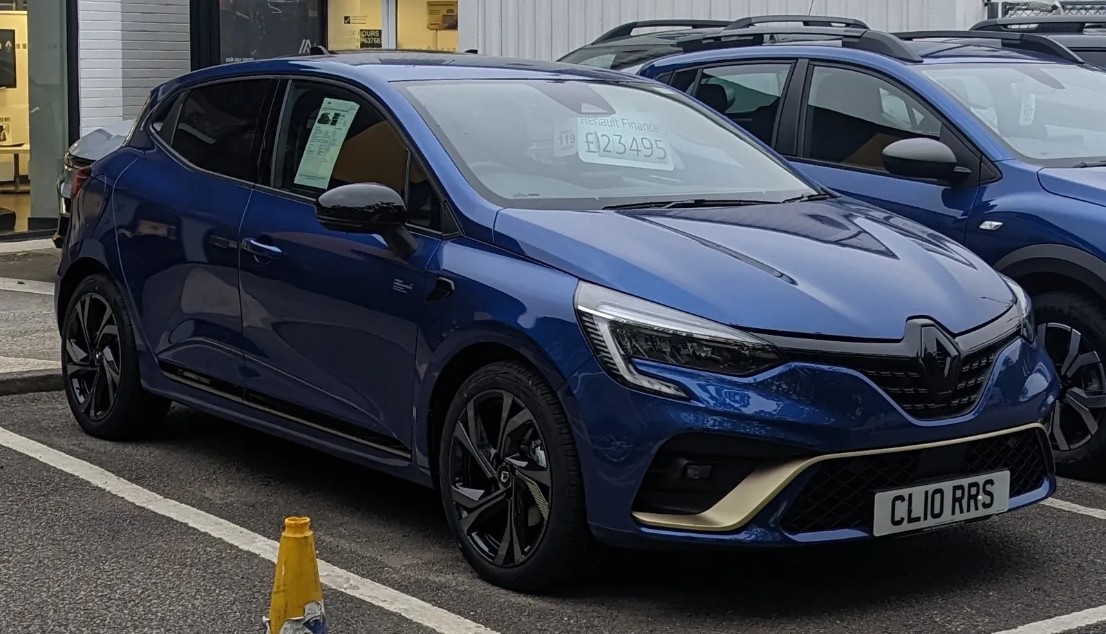 Blue fifth-generation Renault Clio in RS Line trim, photographed from the front three-quarter angle in a dealer forecourt