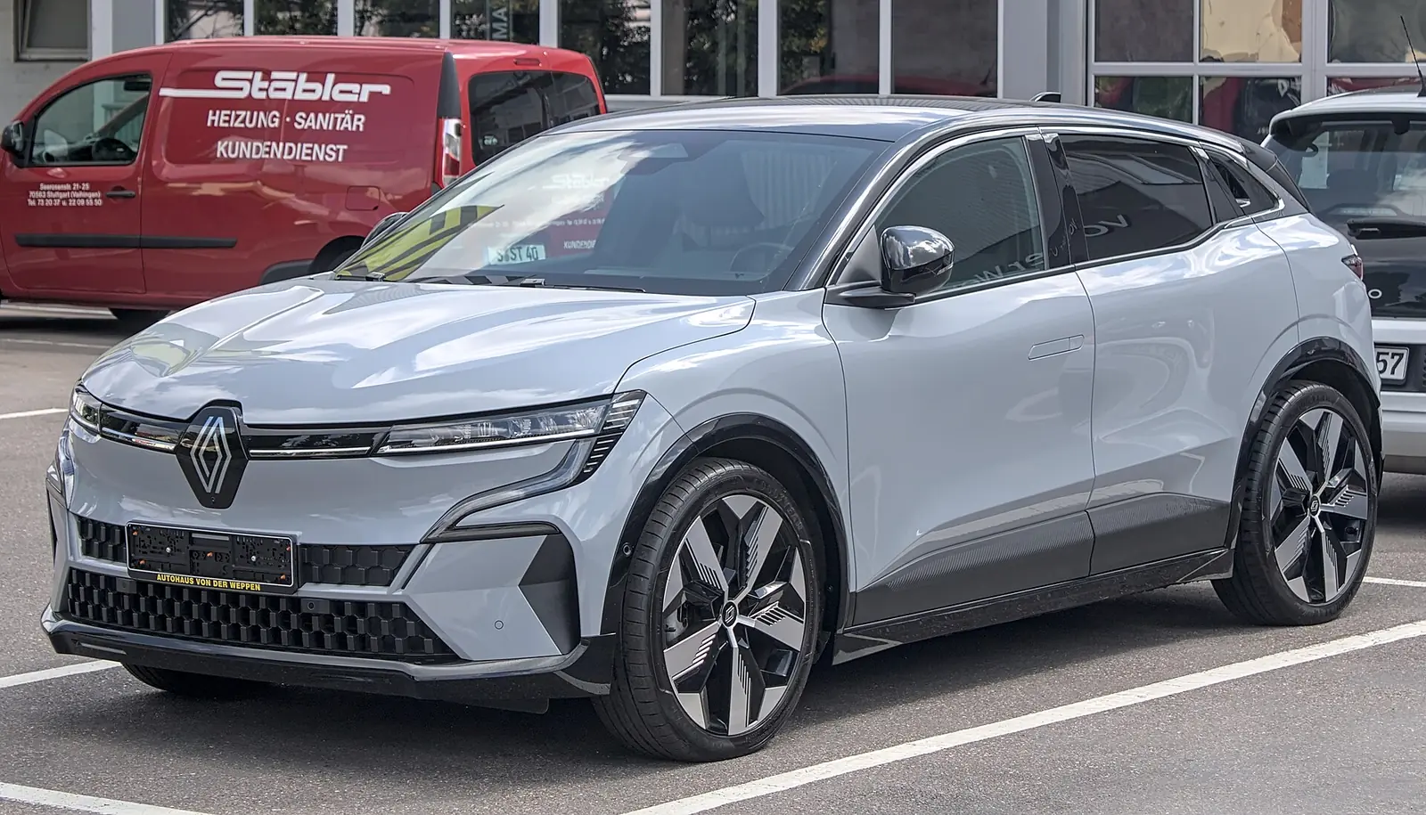 Grey Renault Mégane E-Tech Electric photographed from the front three-quarter angle in a car park, showing its lit diamond-shaped daytime running lights