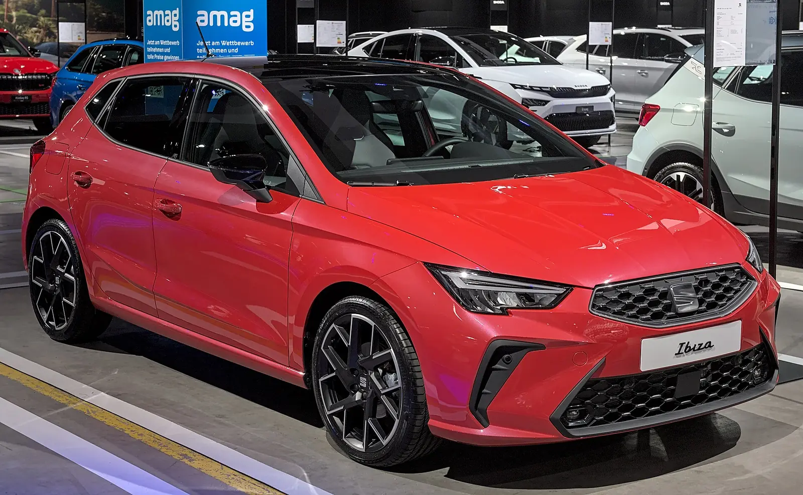 Red current-generation SEAT Ibiza FR photographed from the front three-quarter angle at a motor show, showing the 2025 facelift's hexagonal grille