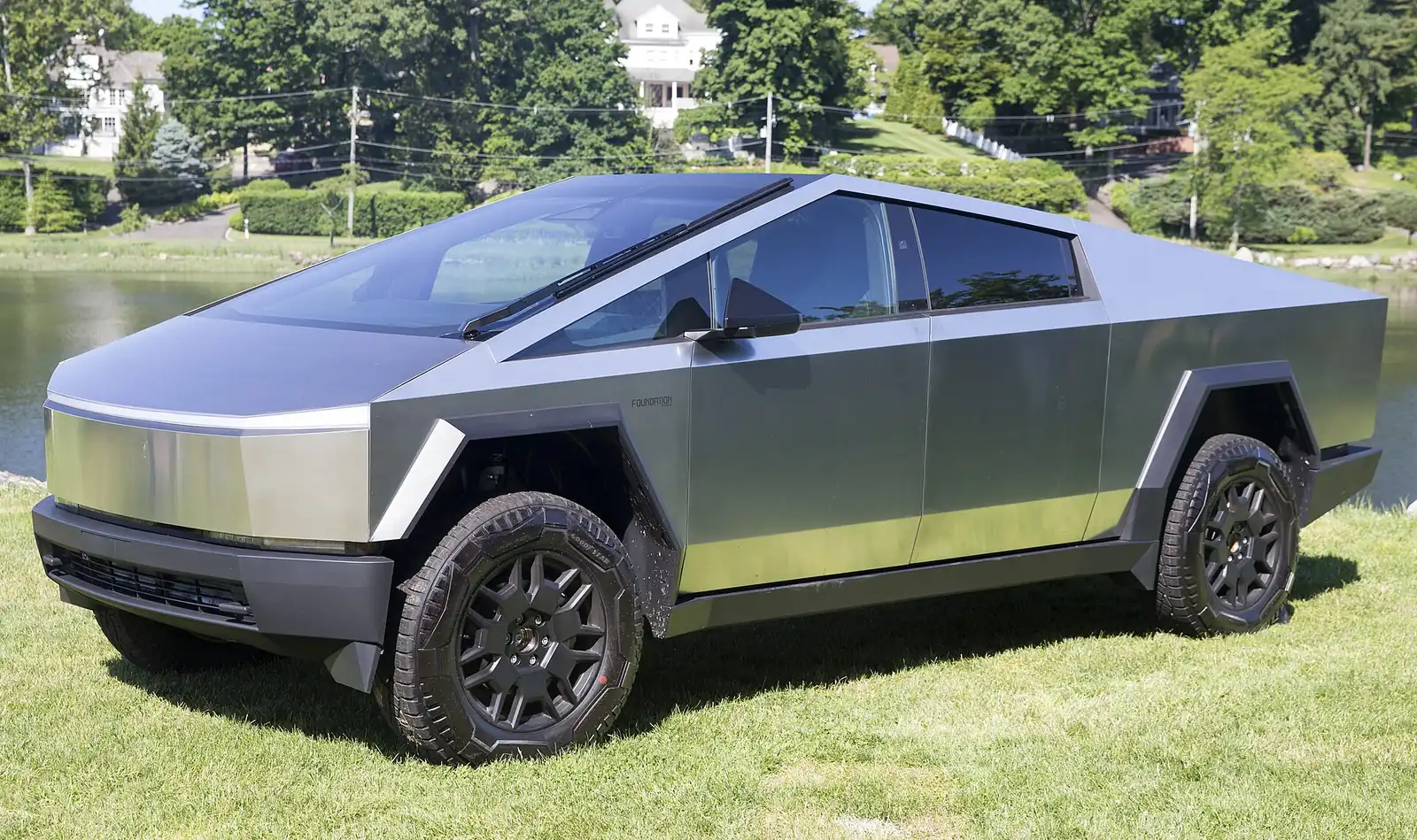 Silver Tesla Cybertruck with angular, faceted stainless-steel bodywork, photographed from the front three-quarter angle on grass beside a lake
