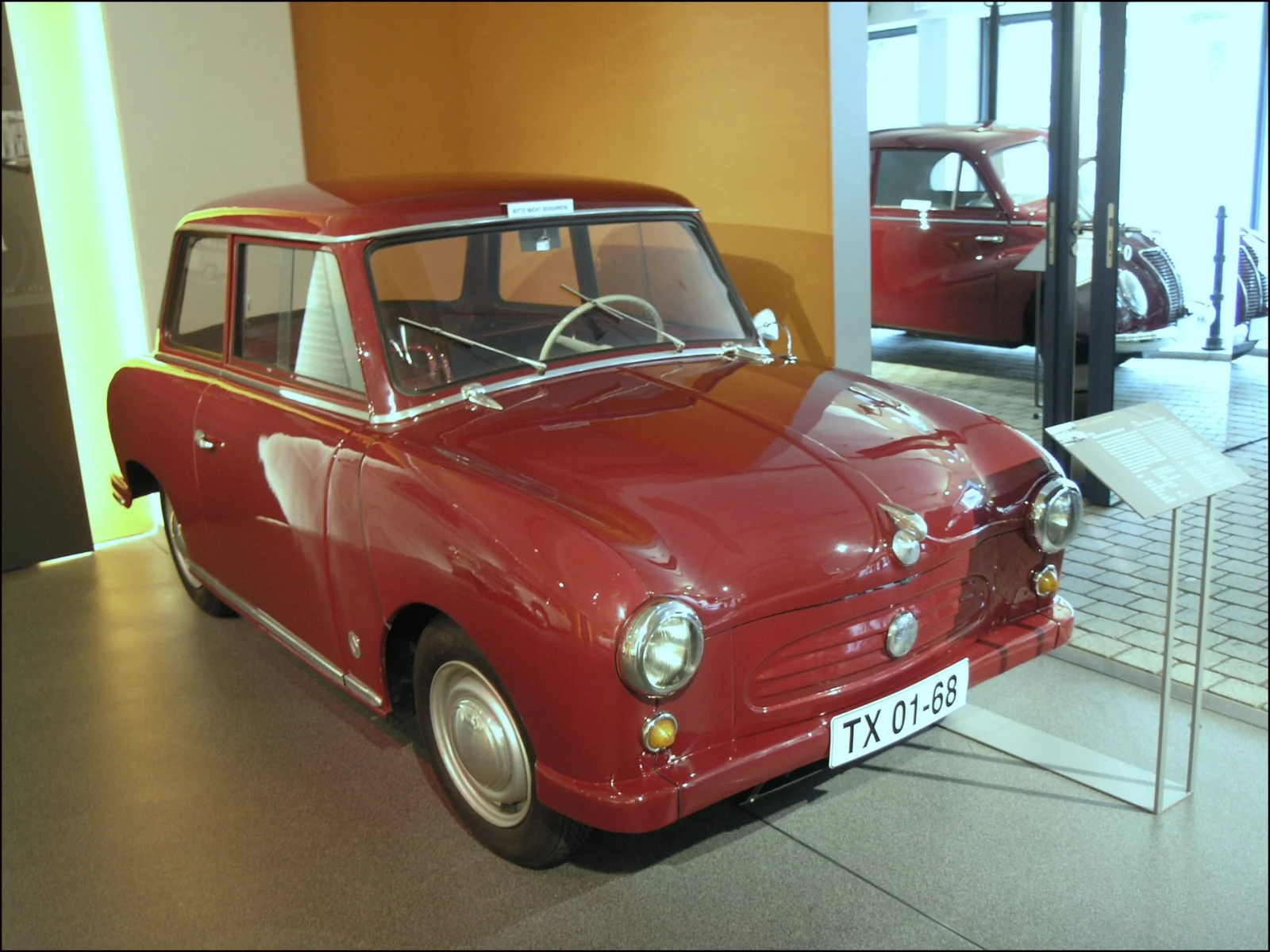 Dark red Trabant P50 photographed from the front three-quarter angle in a museum, showing its rounded 1950s body and single-piece curved windscreen