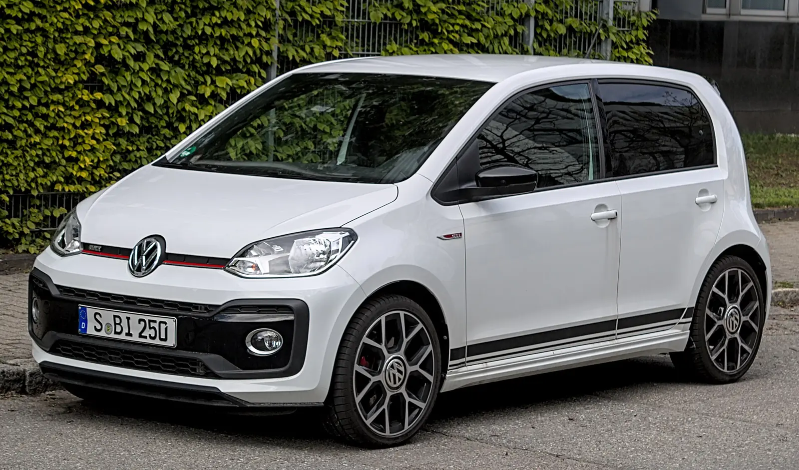 White Volkswagen Up GTI hot hatch photographed from the front three-quarter angle in front of a hedge, with red grille trim and black alloy wheels