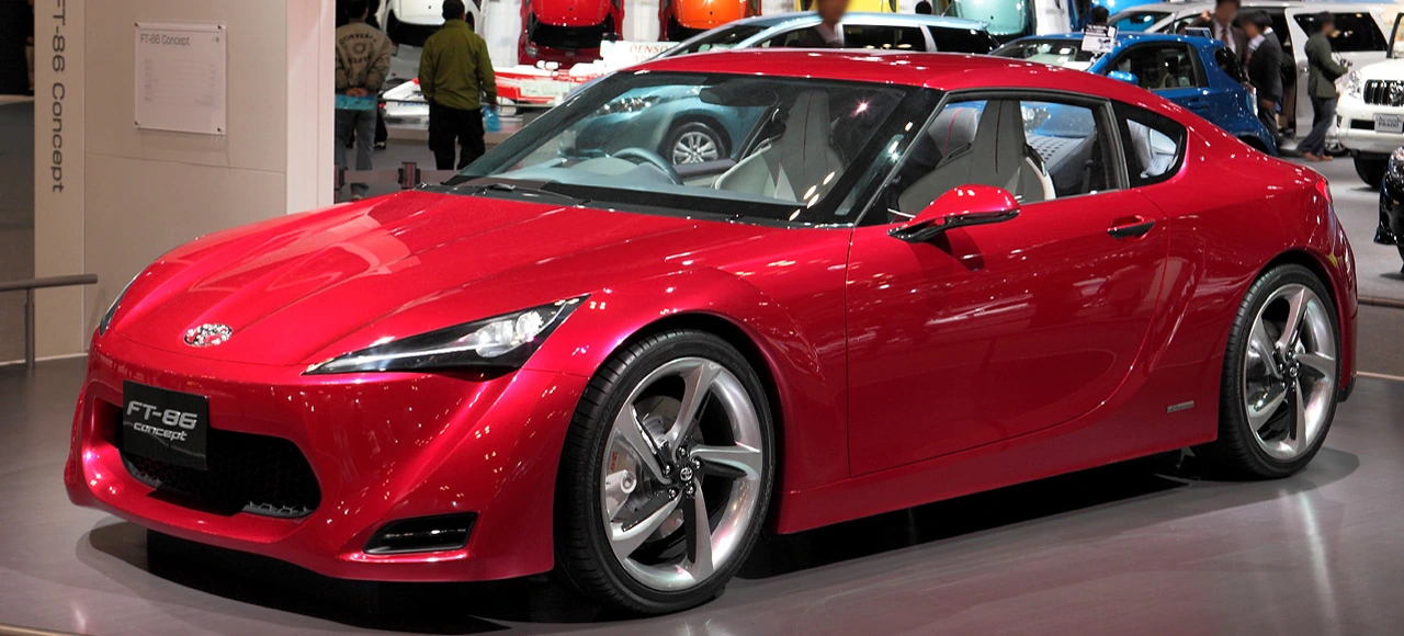 Red Toyota FT-86 Concept coupé on a motor-show stand, seen from the front three-quarter angle, with a low nose and heavily flared wheel arches