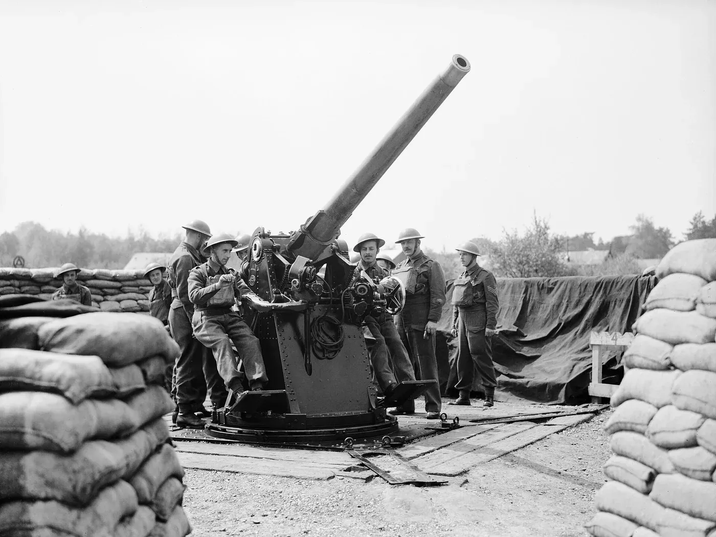 A crew of soldiers in helmets operating a large anti-aircraft gun mounted on a circular platform ringed by sandbags, its barrel angled steeply toward the sky.