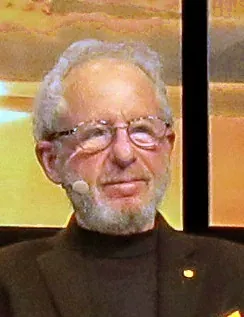 A balding older man with glasses, a moustache and short beard, wearing a headset microphone and a dark turtleneck, photographed at an event.