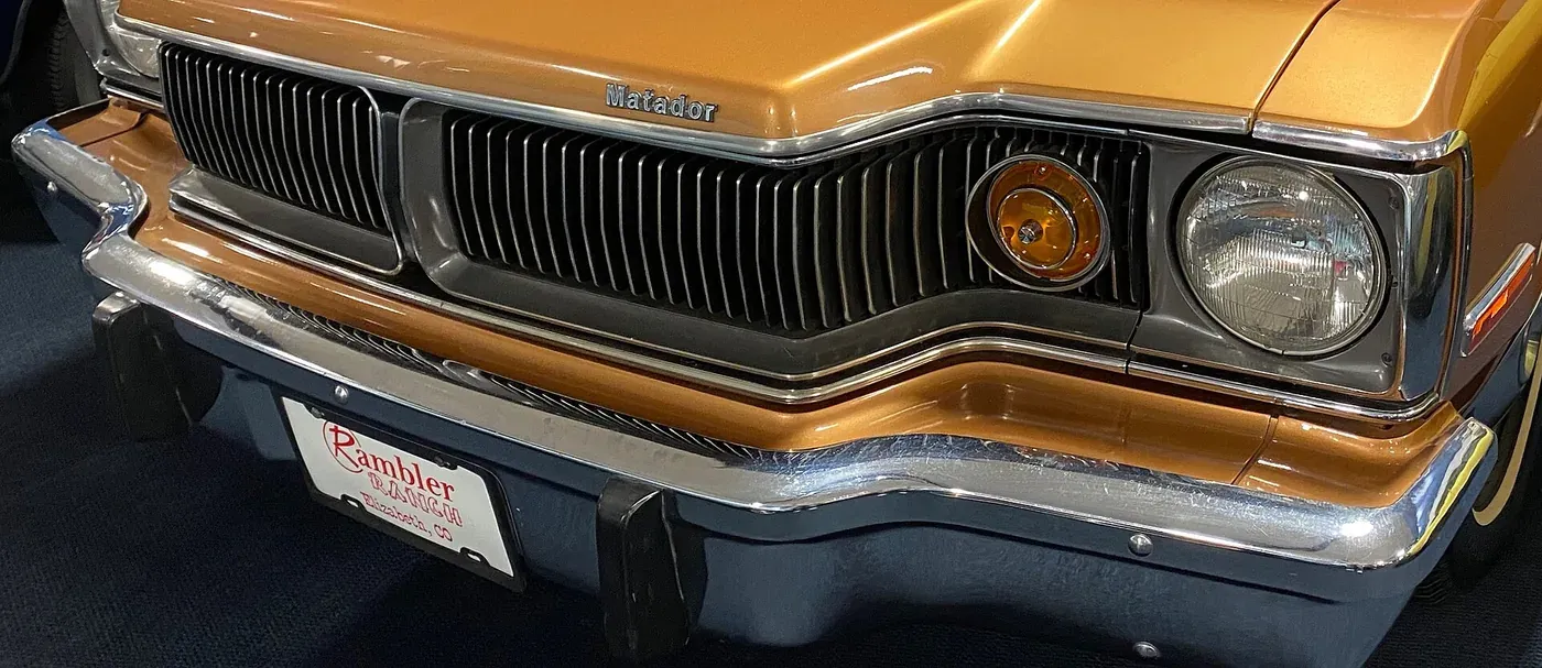Close-up of the front bumper and grille of a glossy tan 1974 AMC Matador, showing a chrome strip over a moulded elastic bumper fascia and a Rambler Ranch dealer plate frame.