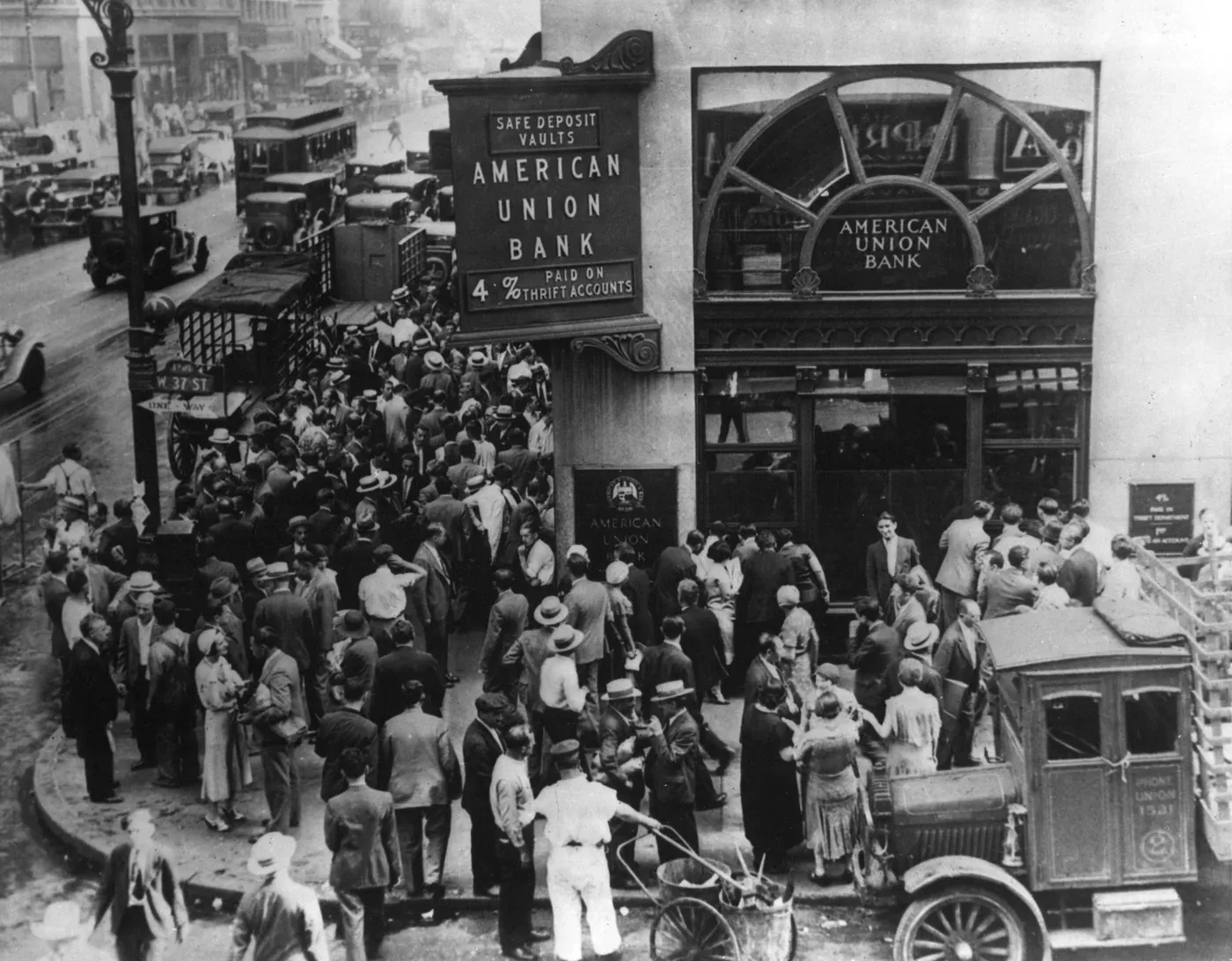 A crowd of people filling a New York City street outside a bank building with a sign reading American Union Bank, seen from an elevated angle, with cars and streetcars backed up alongside.