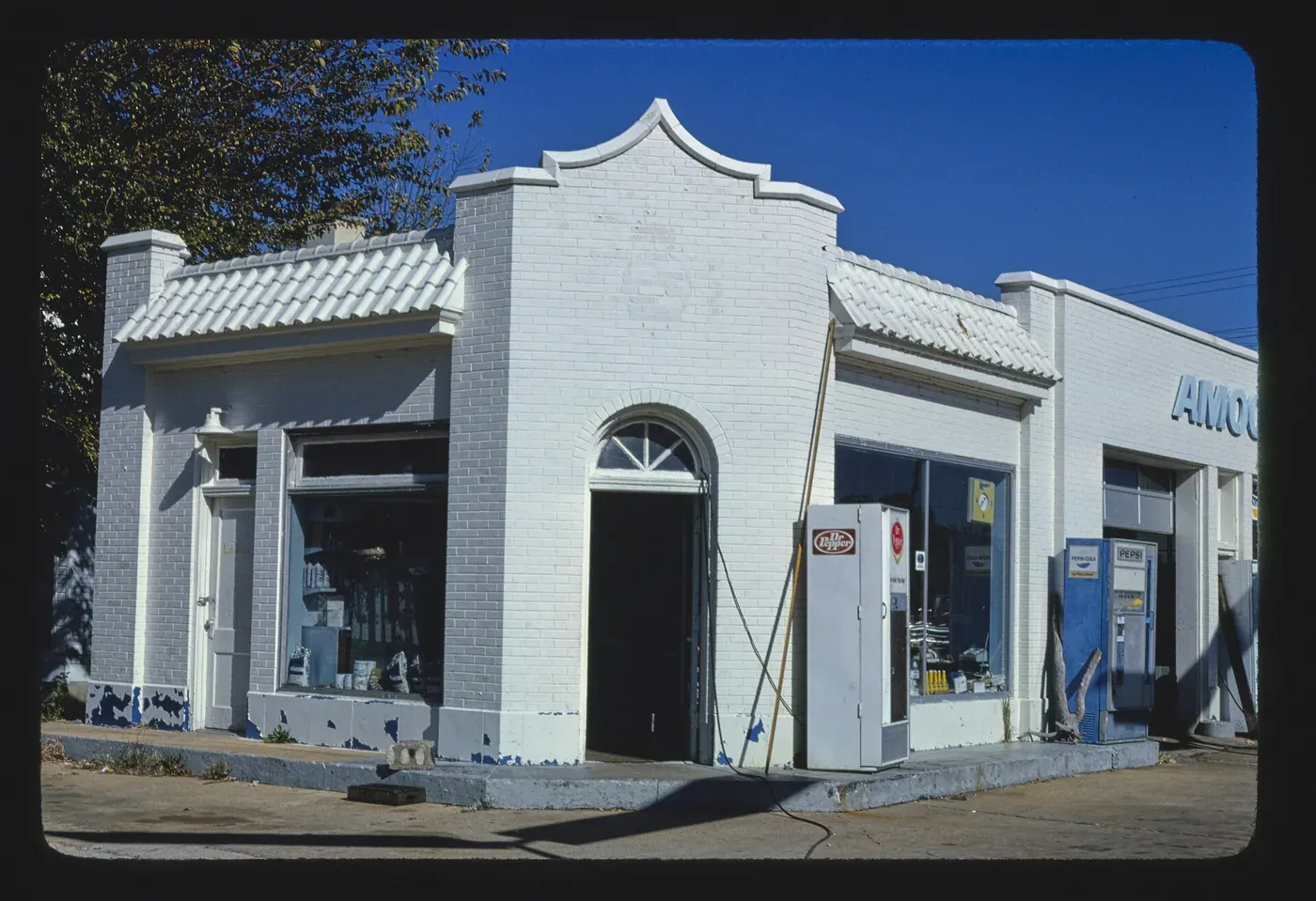 A whitewashed 1920s-style brick filling station with tiled eaves and soda vending machines out front, a partial AMOCO sign visible on the adjoining building, photographed in color.