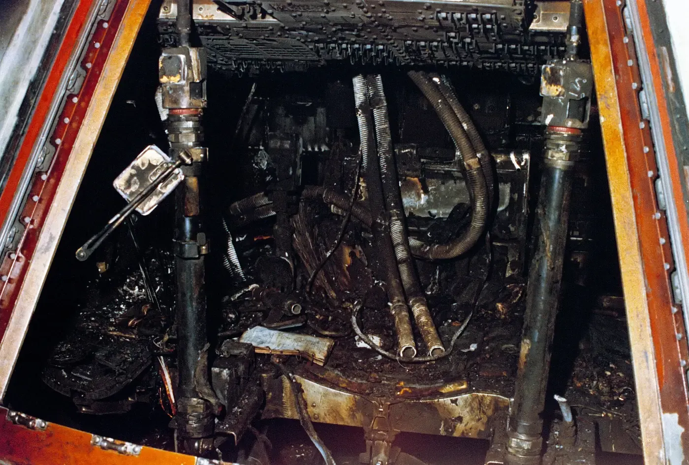 A close view into the scorched interior of a spacecraft command module, its wiring bundles, hoses, and metal fittings charred black and coated in soot.