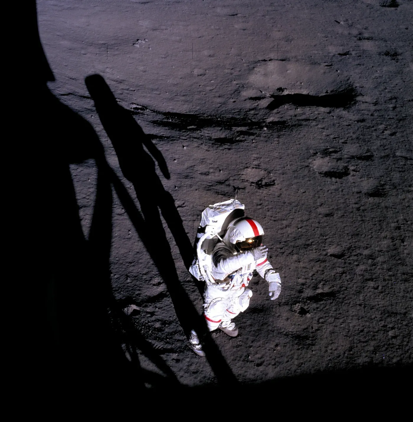 An overhead photograph of an astronaut in a white spacesuit on the grey lunar surface, his helmet's gold-coated visor glinting brightly in direct sunlight, with the dark shadow of a lunar module leg crossing the foreground.