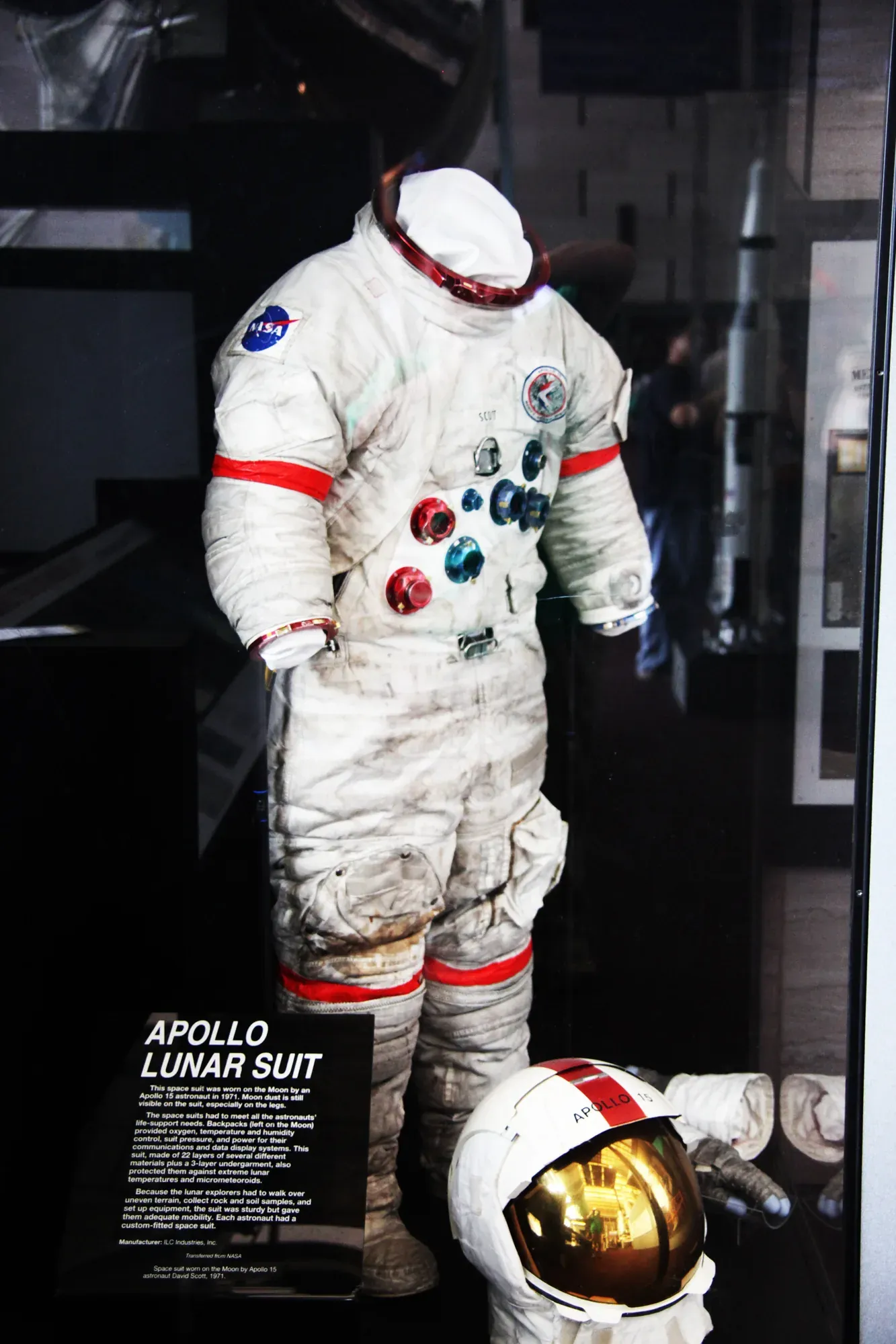A dust-streaked white spacesuit and gold-visored helmet displayed in a museum case, NASA and mission patches on the chest and shoulder, with a placard identifying it as an Apollo lunar suit.