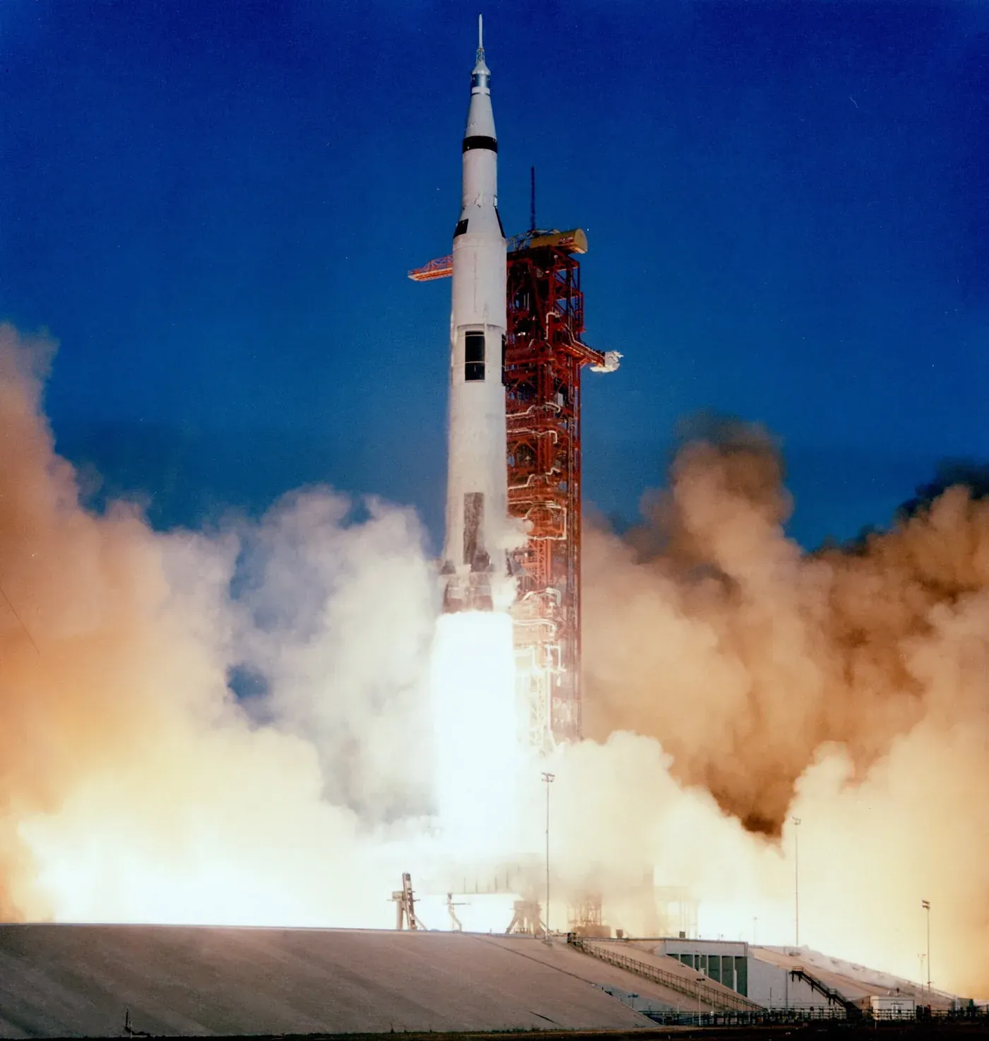 A Saturn V rocket lifting off from its launch pad at night, trailing a huge column of white exhaust and lit orange by its own engine flame.