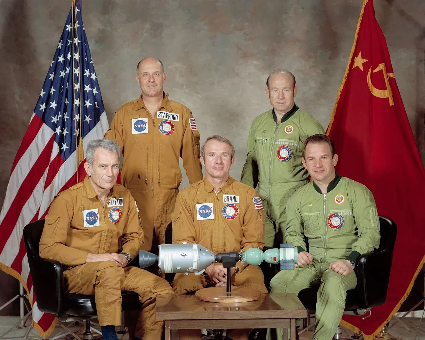 Five men in flight suits pose for a formal portrait, three in tan NASA suits and two in olive Soviet suits, seated around a table holding a scale model of the docked Apollo and Soyuz spacecraft, flanked by American and Soviet flags.