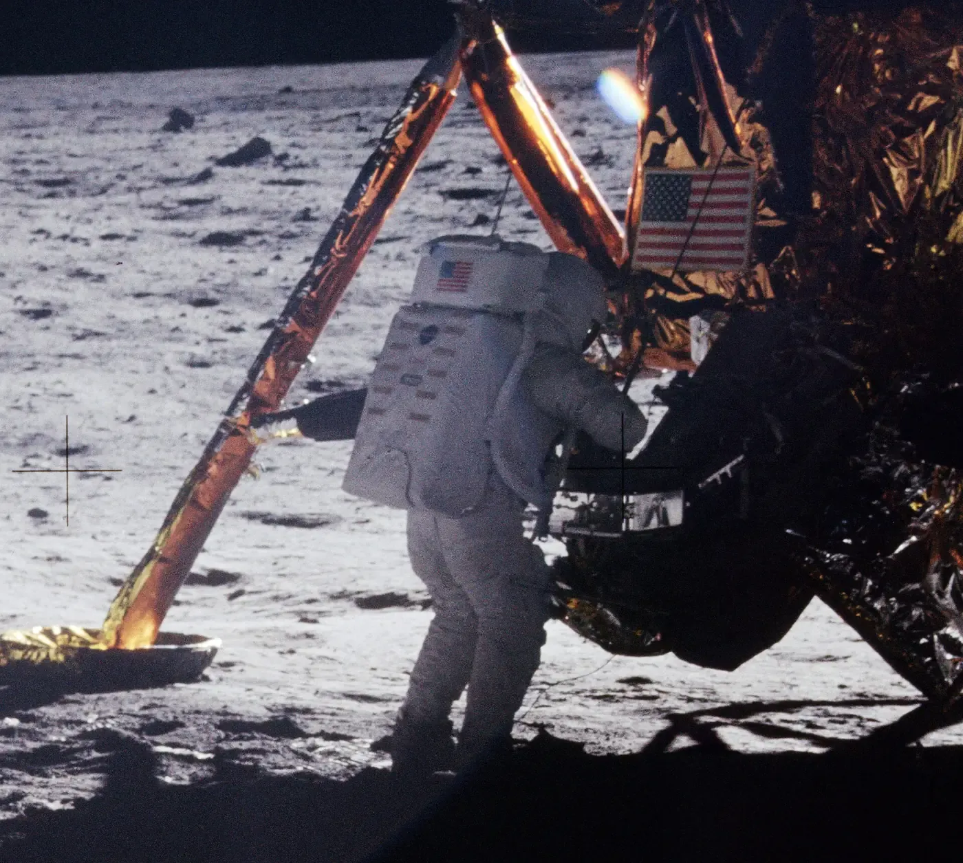 An astronaut in a white spacesuit working beside the gold-foil-wrapped leg of a lunar module on the grey, cratered surface of the Moon, an American flag patch visible on the strut.