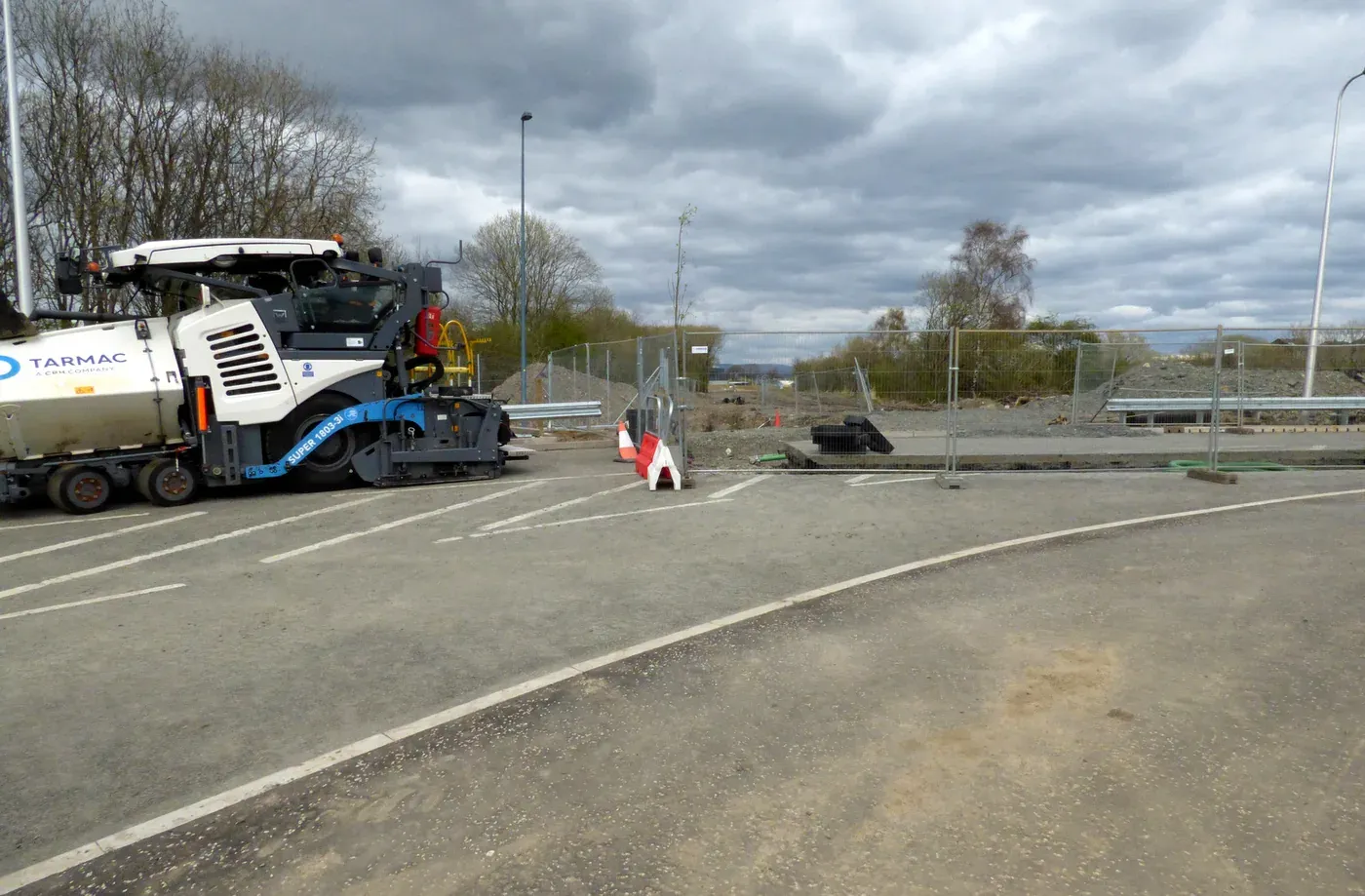 A large asphalt paving machine laying fresh grey asphalt in a car park, under a cloudy sky, with a traffic cone and safety fencing nearby.