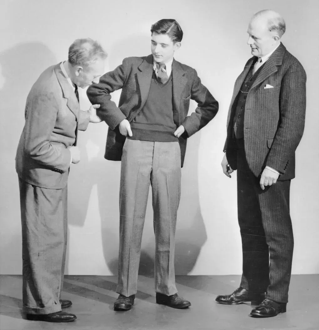 Three men in a photography studio: one in the centre wears a jacket and trousers from the wartime utility clothing scheme while an older man beside him inspects the sleeve, and a third man in pinstripe suit looks on.