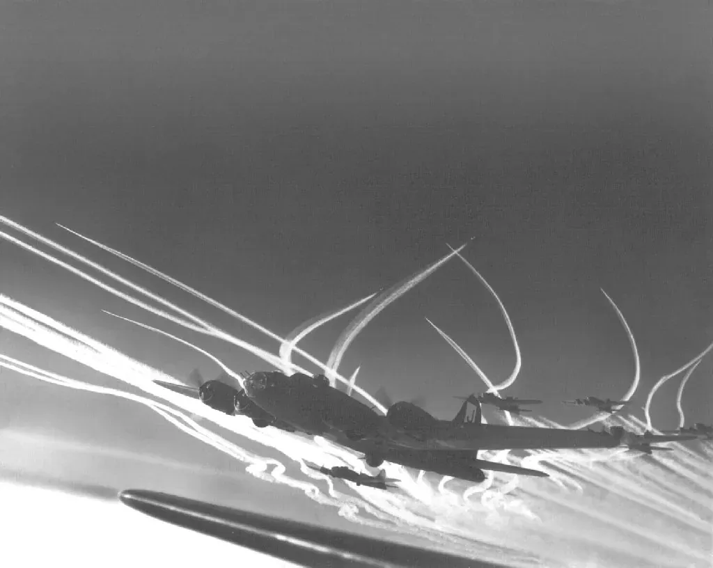 A formation of B-17 bombers and P-47 fighter escorts seen from below, trailing long white vapor contrails against a dark sky.