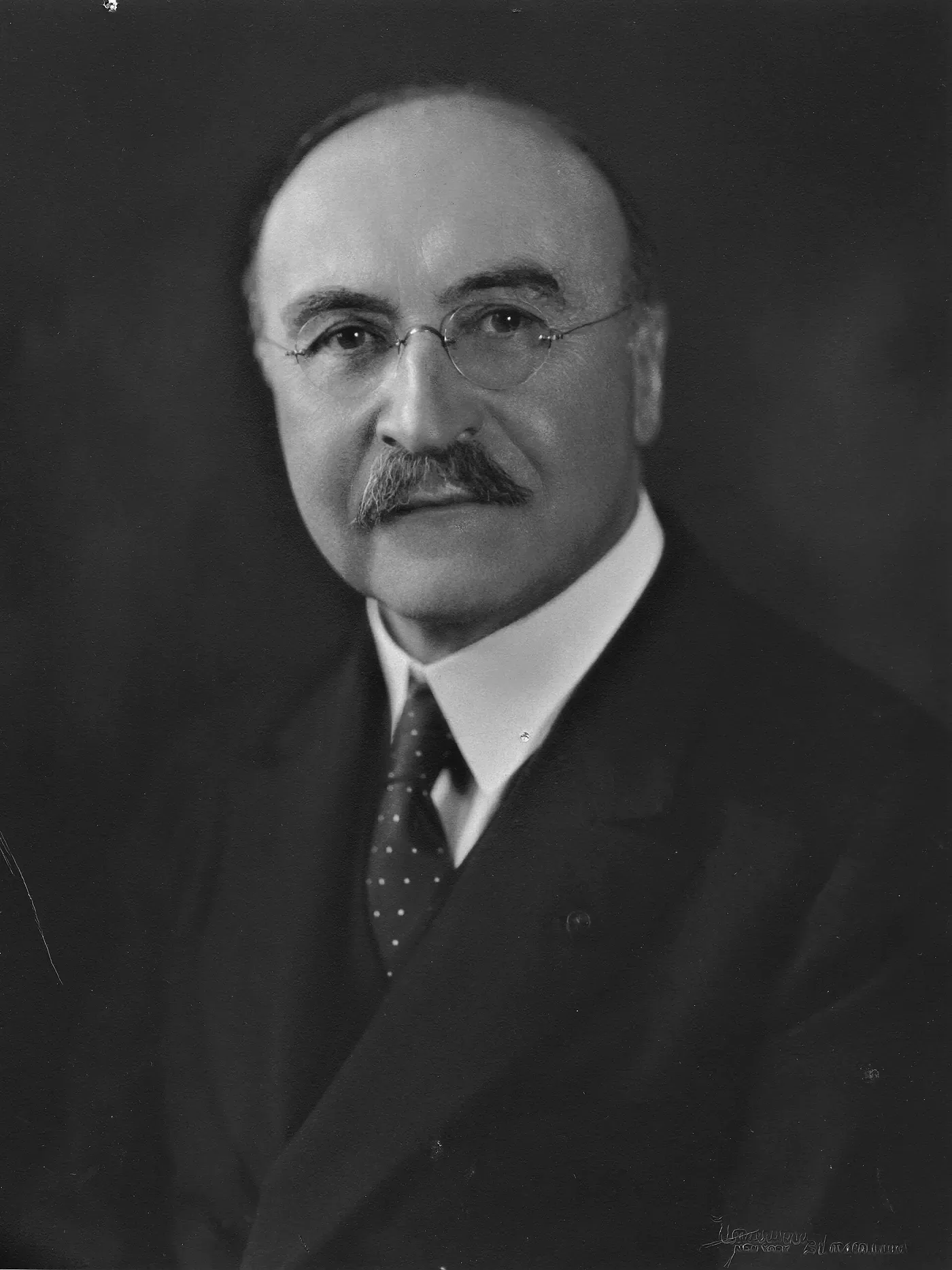Studio portrait of Leo Baekeland in a dark suit, balding, with a heavy moustache and wire-rimmed spectacles.