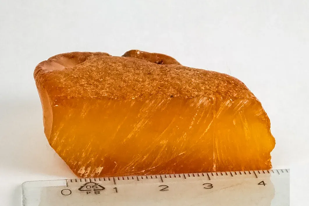 A rough, translucent orange-gold lump of raw amber, its cut face showing streaky internal patterning, resting beside a clear plastic ruler for scale.