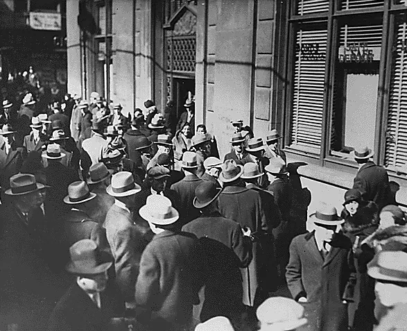 A dense crowd of men and women in coats and hats gathered on a city sidewalk outside a bank building with shuttered windows.