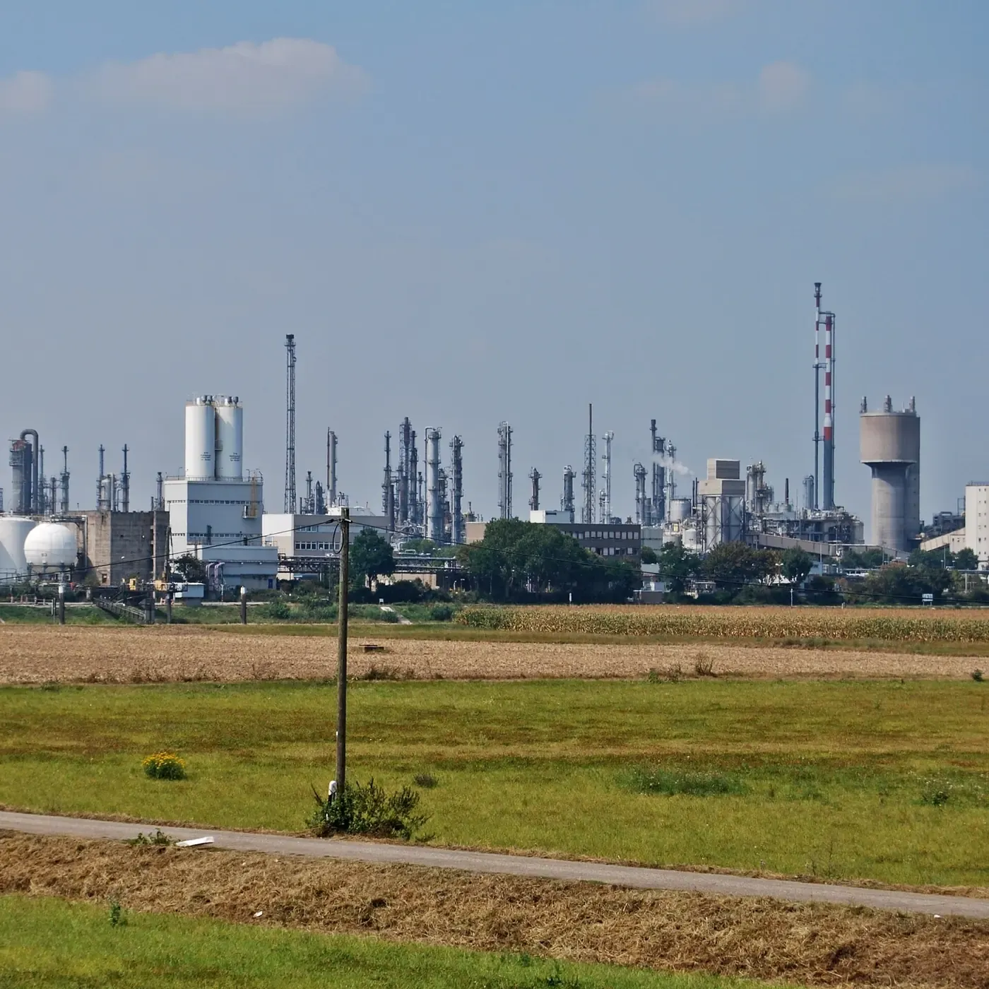 A wide view across farmland toward a large chemical plant on the horizon, its skyline dense with distillation towers, storage tanks and a tall red-and-white striped chimney.
