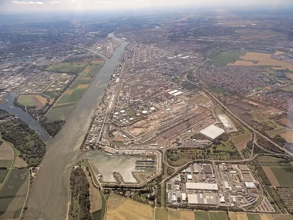 An aerial photograph looking down a wide river bend crowded on one bank with dense industrial plant (storage tanks, pipe racks and processing units) surrounded by farmland.