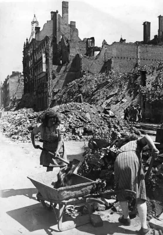 Two women in aprons and headscarves shoveling rubble into a wheelbarrow in front of a bombed, roofless building, with a large mound of broken masonry behind them.