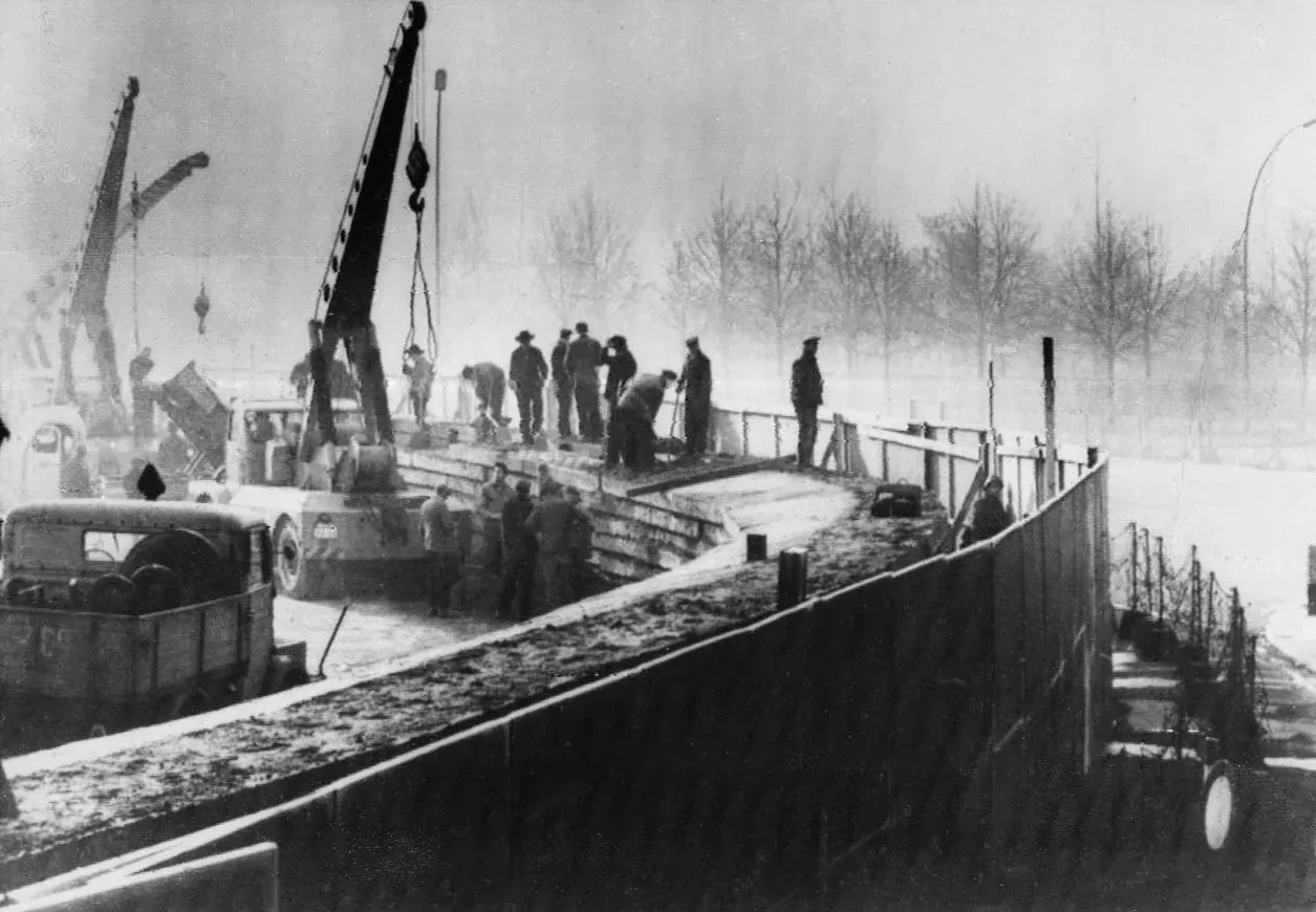 Black-and-white photograph of construction workers and cranes building a low concrete wall topped with barbed wire, on a frosty morning.