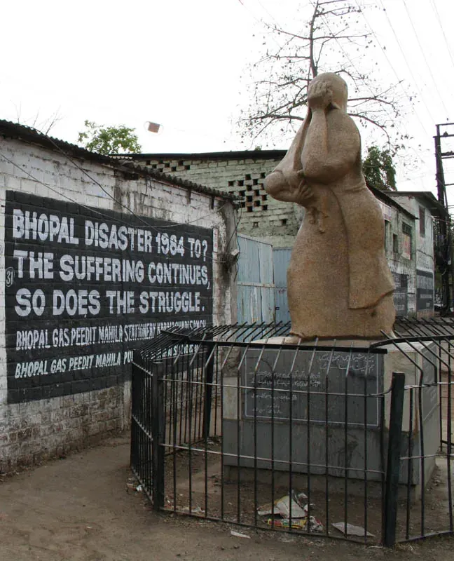 A stone memorial statue of a mourning mother and child behind a metal fence, beside a wall painted with the words BHOPAL DISASTER 1984 TO? THE SUFFERING CONTINUES, SO DOES THE STRUGGLE.