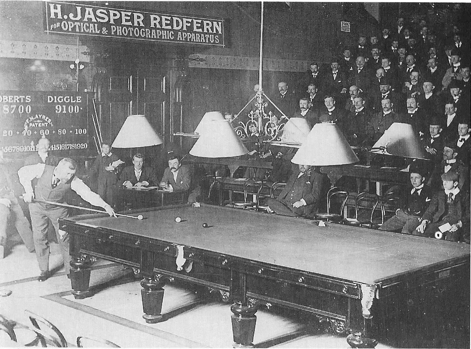 A crowded Victorian billiard hall with two men playing on a large table lit by hanging shaded lamps, watched by rows of seated spectators in formal dress.