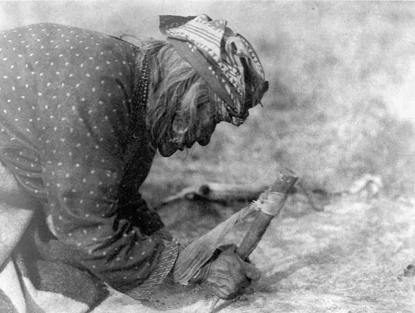 A black-and-white photograph of a woman kneeling outdoors, scraping the flesh side of an animal hide staked to the ground with a bone tool.