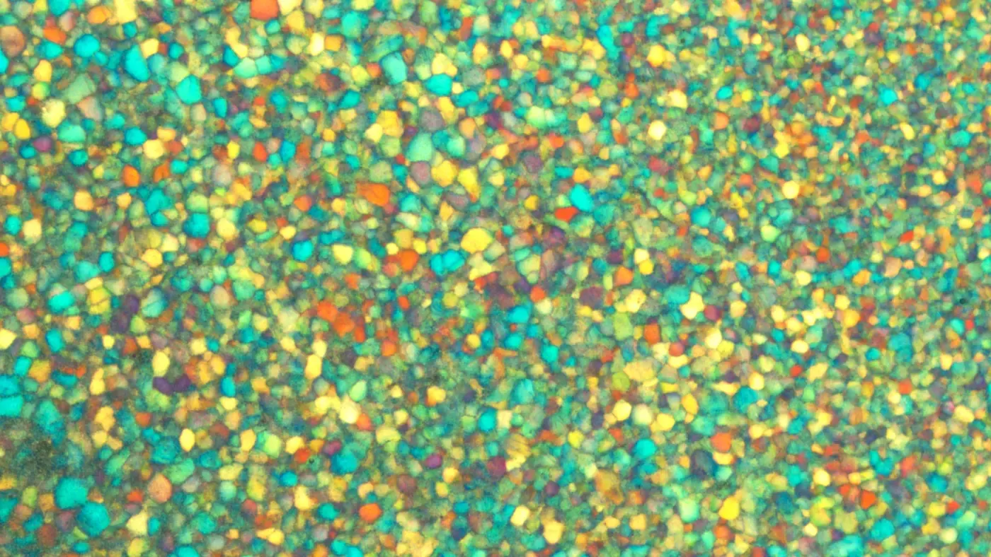 A close-up polarized-microscope image showing a dense, irregular mosaic of small angular crystal platelets in bright teal, yellow, orange and violet.