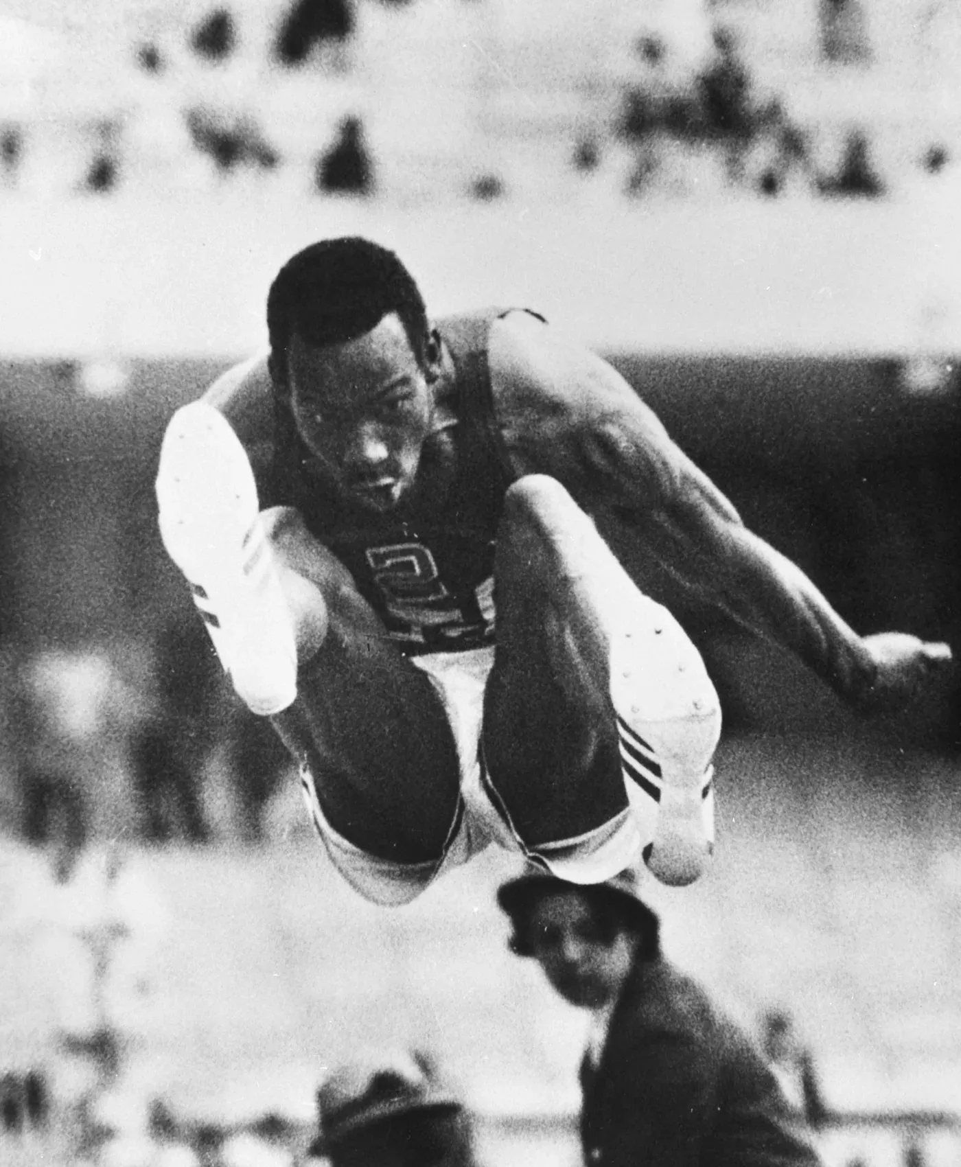 A close-up action photograph of an athlete in mid-air during a long jump, knees drawn up toward his chest, arms trailing behind, with a blurred stadium crowd in the background.