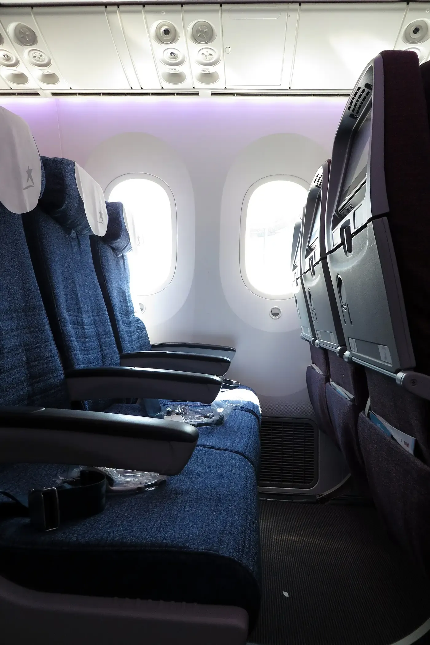 A close-up view inside an aircraft cabin showing two rows of blue upholstered economy seats beside oval windows, with white overhead panelling and ventilation nozzles visible above.