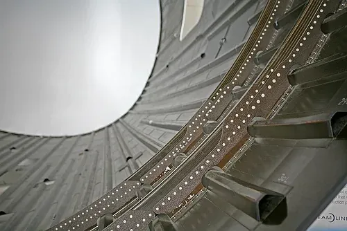 A close-up interior view of a large curved carbon-fiber composite fuselage section, its dark woven surface reinforced along riveted structural frames.