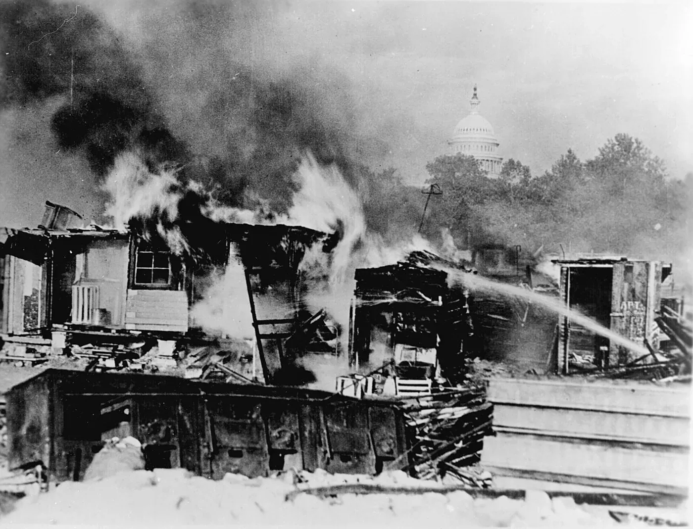 Black-and-white photograph of wooden shacks engulfed in flames and thick smoke, with the U.S. Capitol dome visible through the haze in the background.