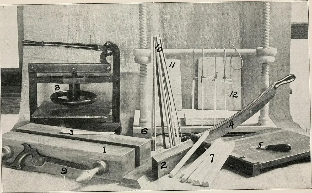 A labelled photograph from a 1914 instructional manual showing a bookbinder's press, sewing frame, folding sticks, paring knife and other hand tools arranged on a table.