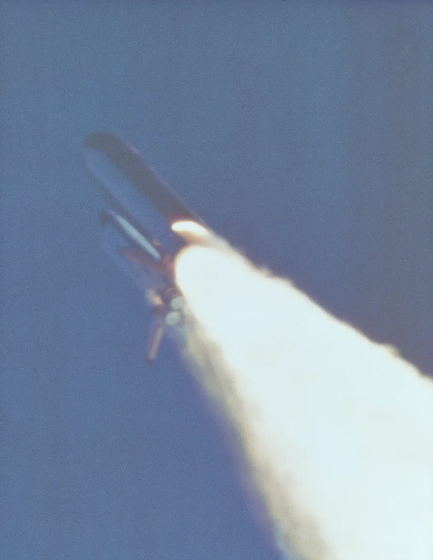 A grainy telephoto frame of the Space Shuttle in flight against blue sky, with a bright orange flame plume jetting sideways from low on the solid rocket booster.
