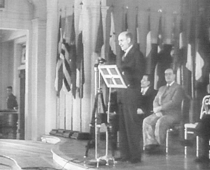 A black-and-white photograph of a man addressing an audience from a lectern on a stage draped with national flags, with two seated men beside him.