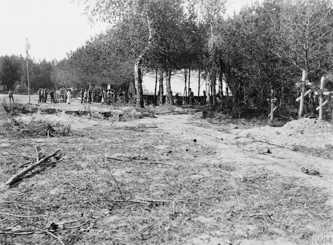 A black-and-white photograph of a wartime camp clearing among birch and pine trees, with a group of soldiers gathered at a tent in the middle distance and wooden crosses marking graves at the right.