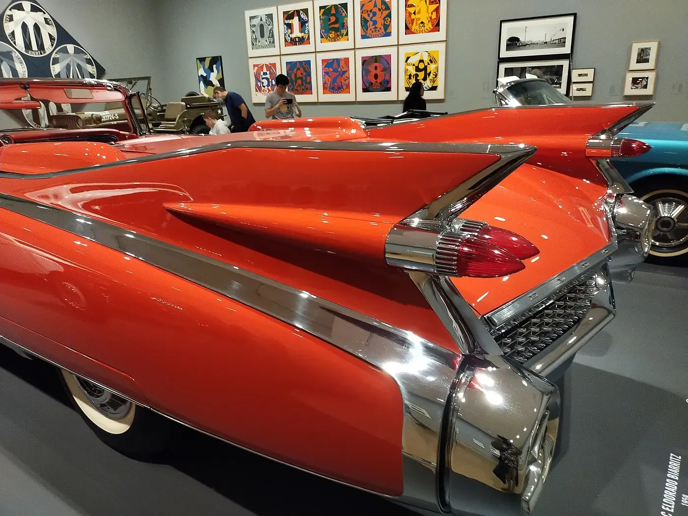 The rear end of a bright orange-red 1959 Cadillac Eldorado, showing its towering chrome-edged tailfins and twin bullet tail-lights, displayed in a museum gallery.