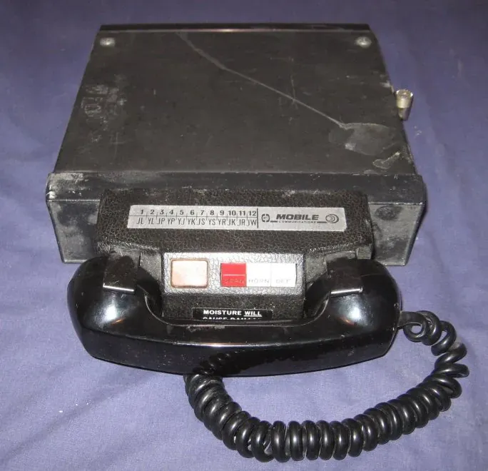 A boxy black car-mounted mobile radio telephone unit with a rotary-style handset, a numbered channel selector strip and a moisture warning label.