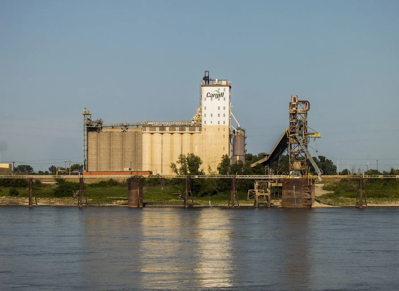 A large concrete grain elevator complex beside a river, with the Cargill company logo painted on its tallest tower and a rail bridge crossing the water in the foreground.