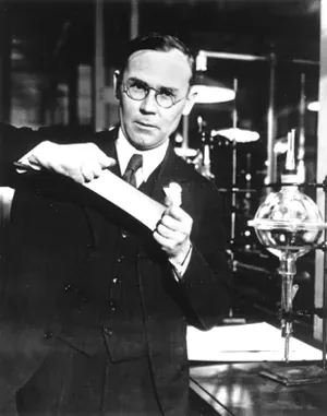 A man in glasses and a suit stands at a laboratory bench holding up a coiled strip of material in one hand and a small sample in the other, with glassware and a Bunsen burner behind him.