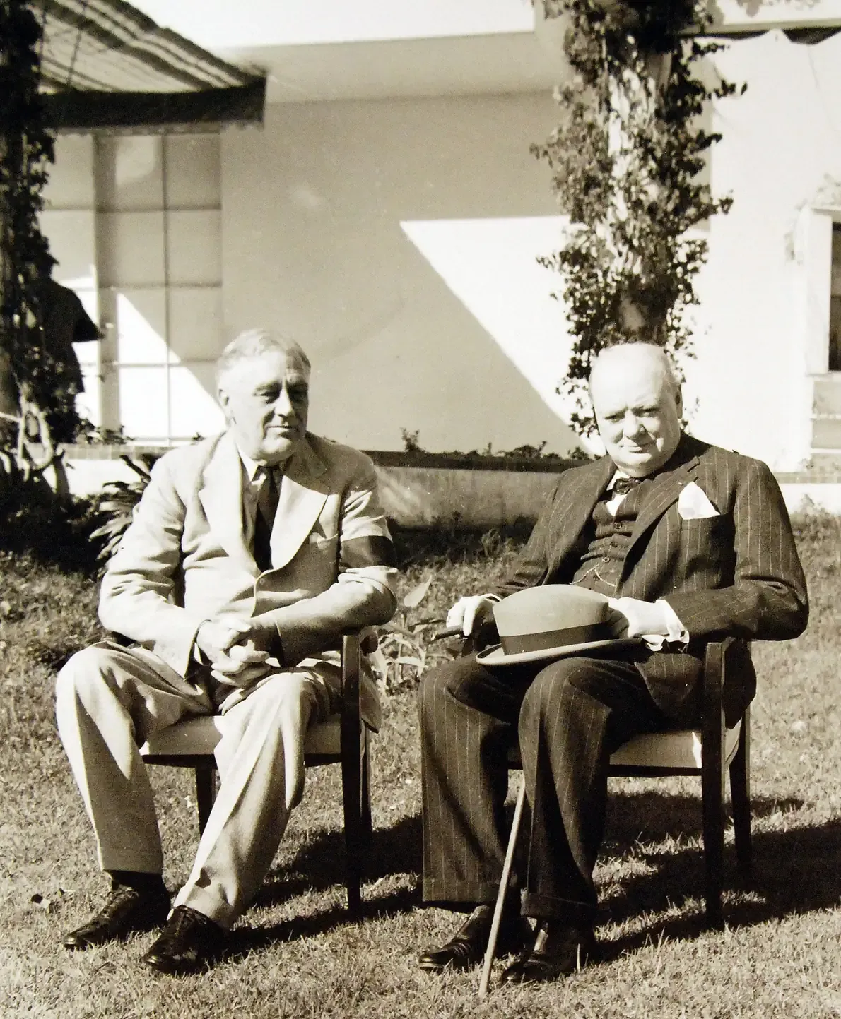 Franklin D. Roosevelt and Winston Churchill seated side by side in wicker chairs on a villa lawn, Churchill holding a hat and cane.